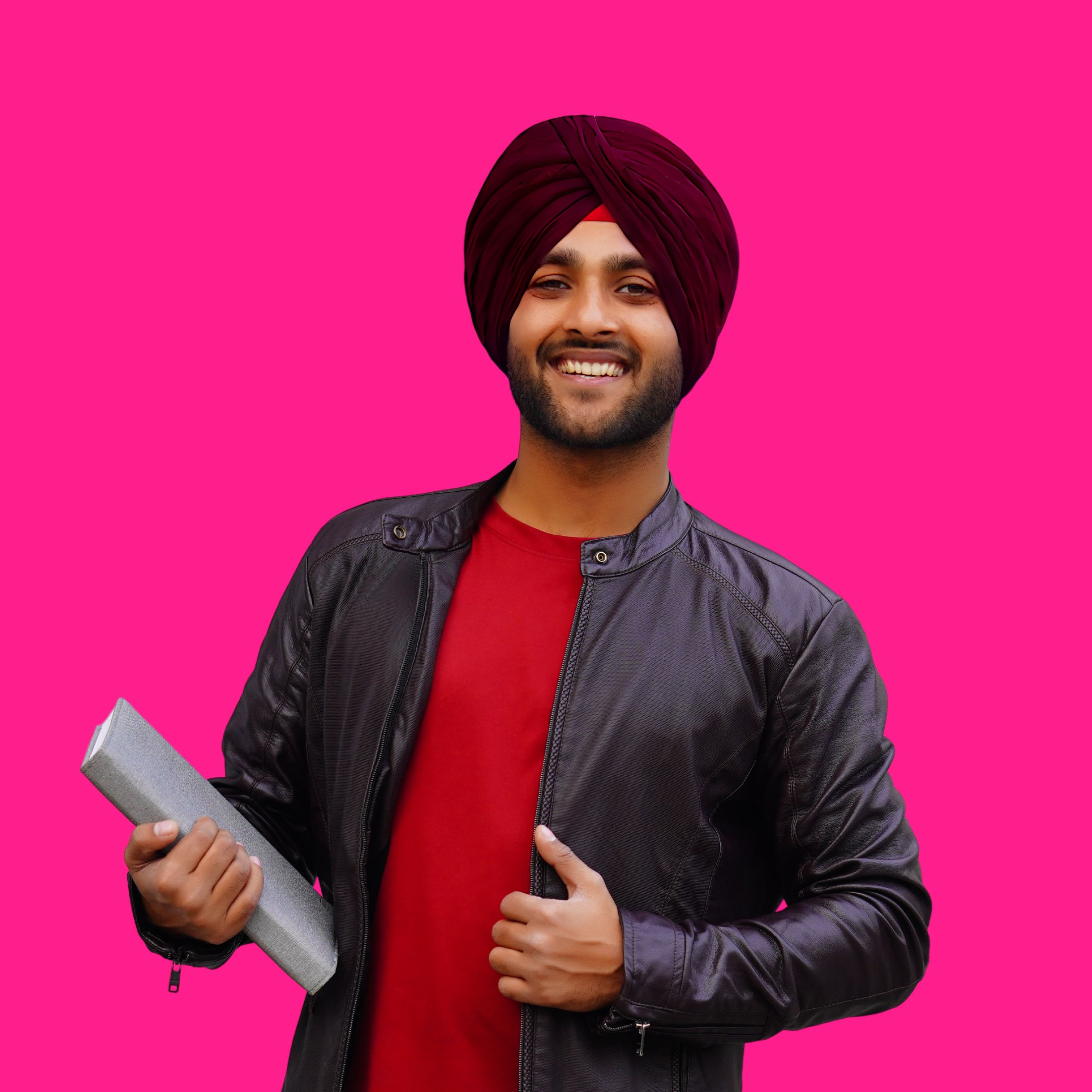 A man wearing a turban, smiling and working on a laptop against a hot pink background