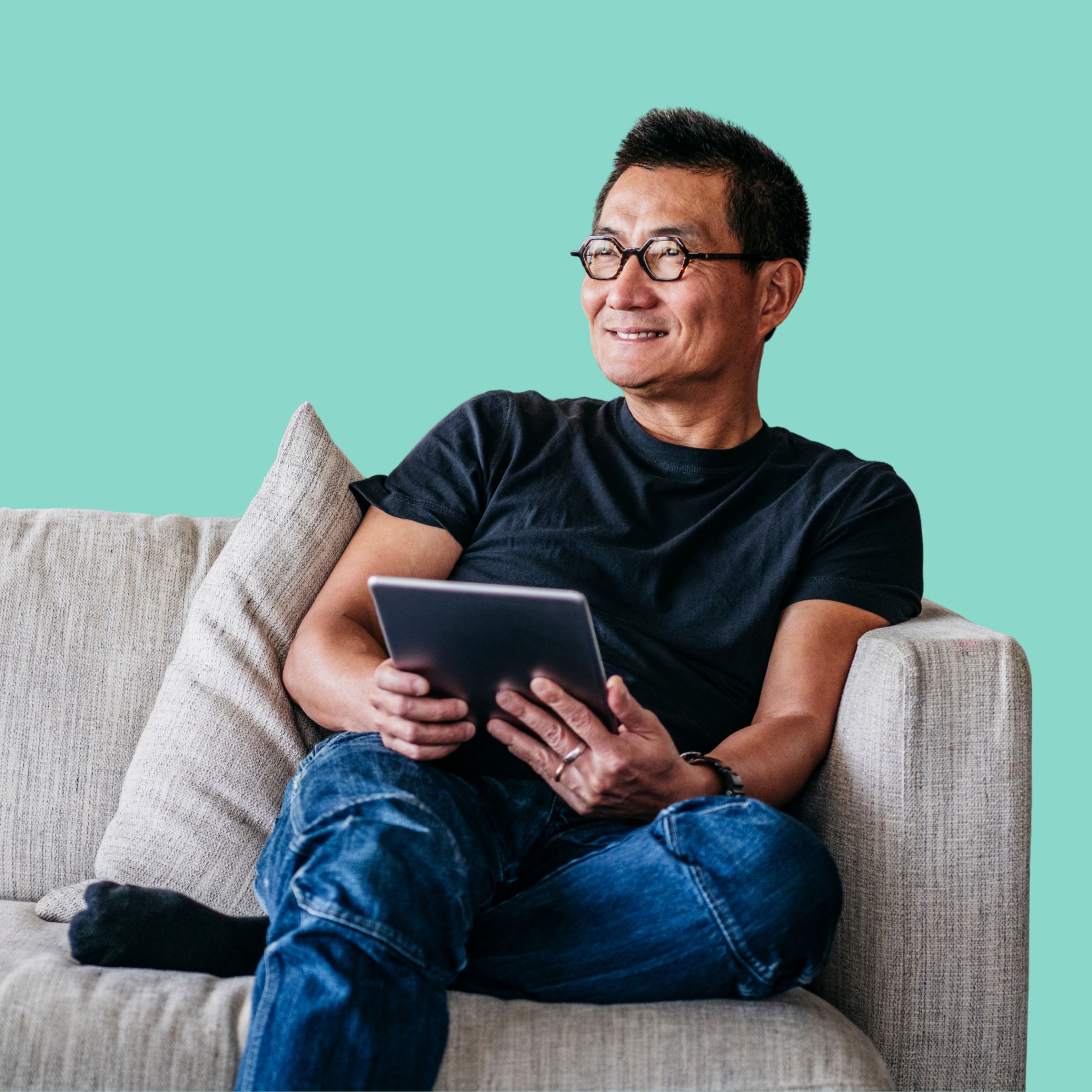 A man wearing glasses sitting on a couch with a tablet, looking thoughtfully into the distance against a turquoise background